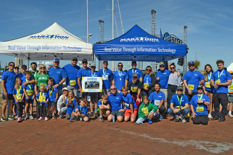 Group photo of Marathon Consulting employees, friends, and family in front of tents in the sun wearing blue Marathon tshirts